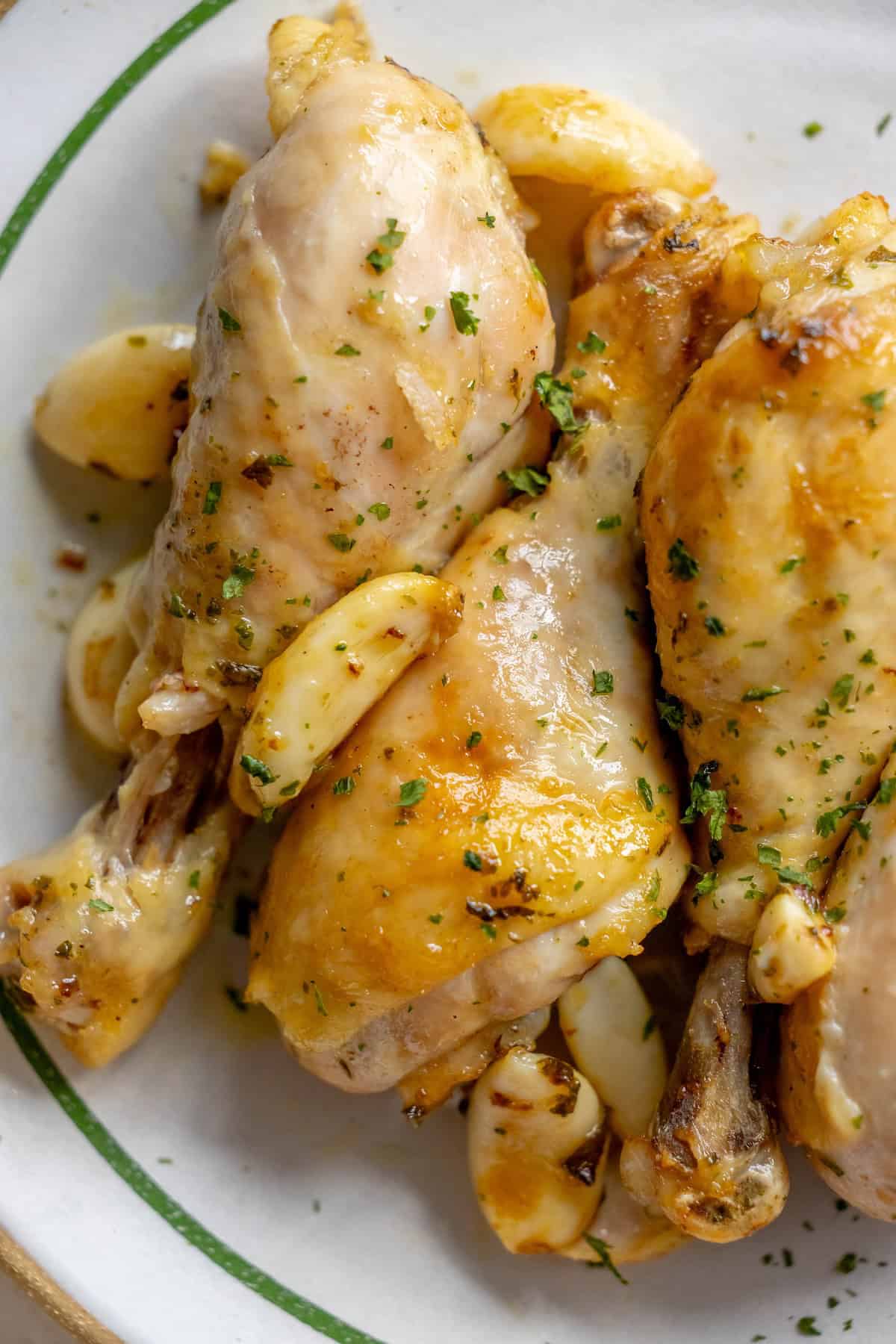 Close-up of Greek chicken drumsticks with roasted garlic cloves and herbs on a white dish.