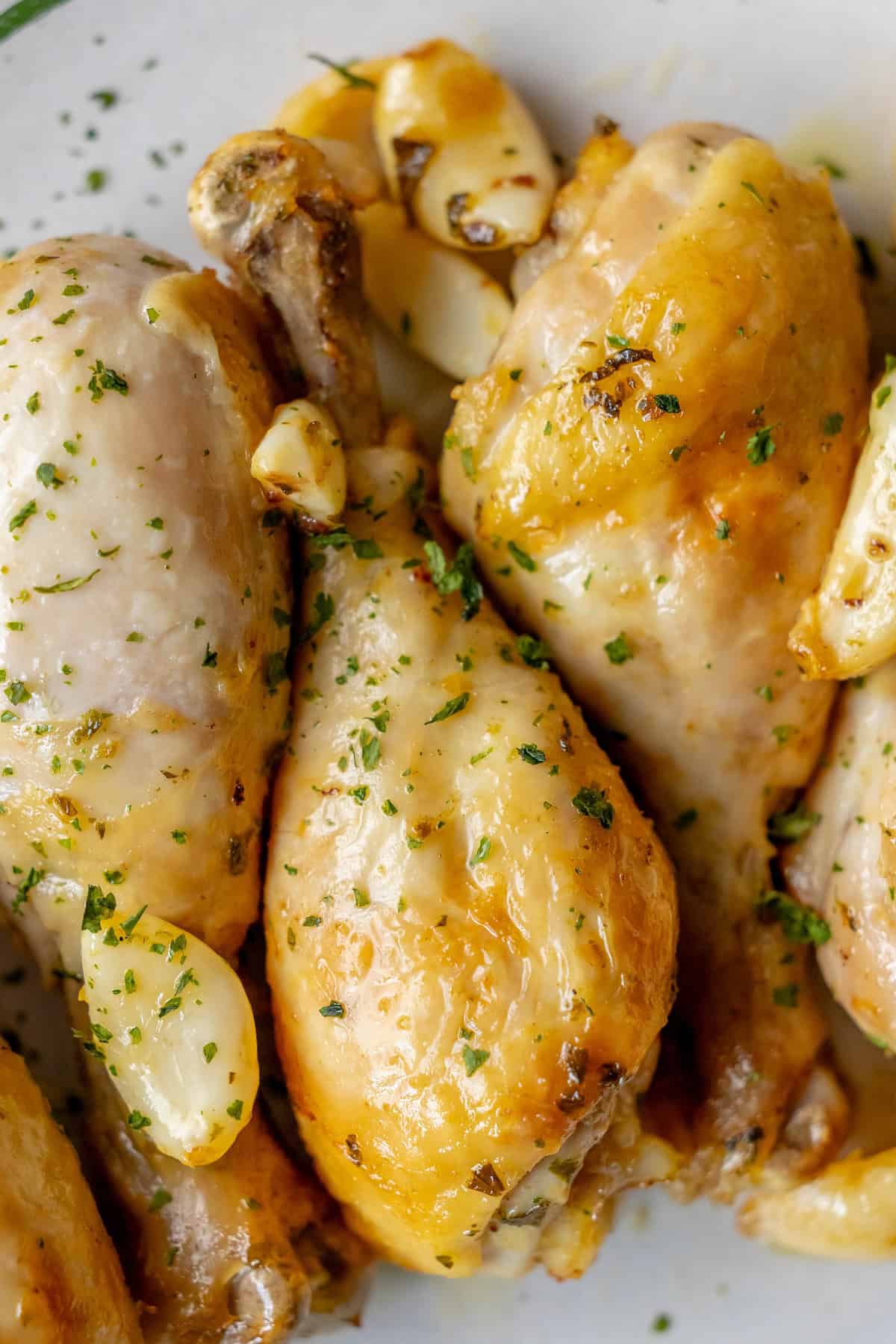 Close-up of Greek chicken drumsticks, roasted to perfection and garnished with chopped herbs and garlic cloves on a white plate.