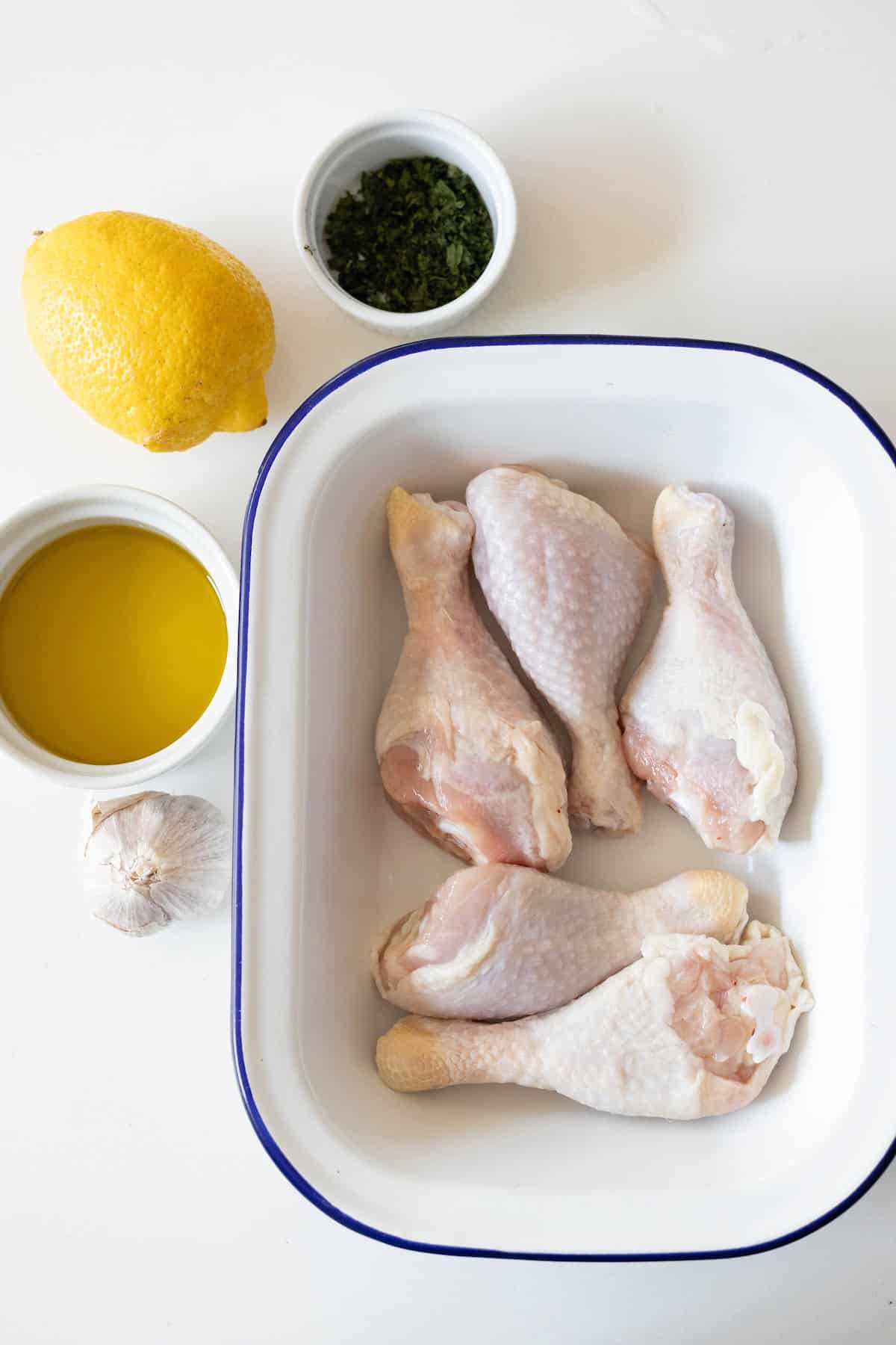 Greek Chicken Drumsticks nestle in a white dish, surrounded by a lemon, a bowl of olive oil, garlic, and a small cup of chopped herbs on a pristine white surface.
