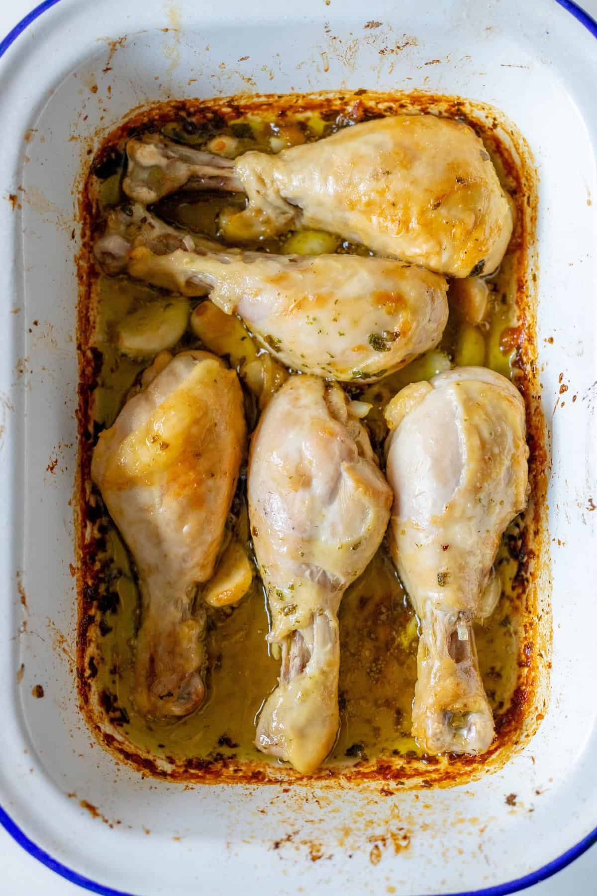 Five Greek chicken drumsticks, perfectly roasted, rest in a white baking dish.