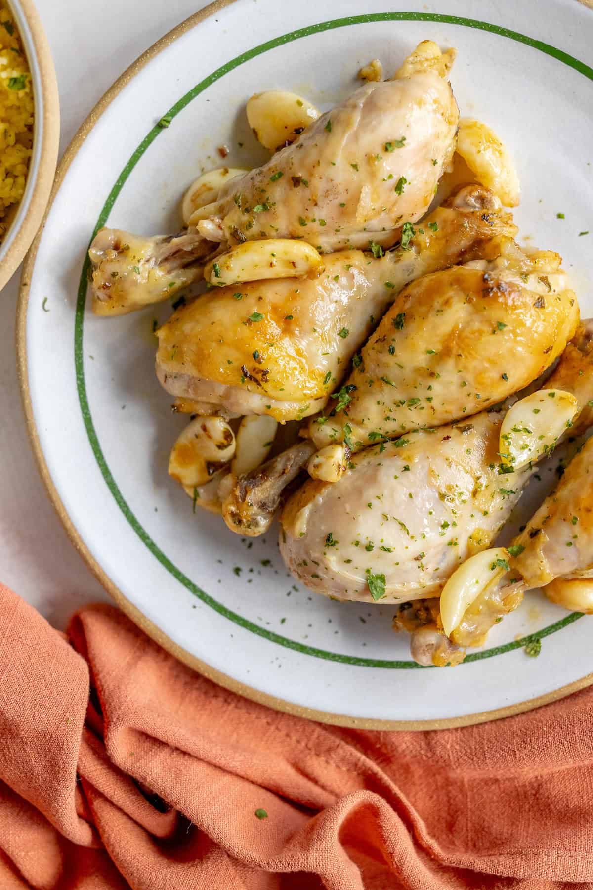 A plate of Greek chicken drumsticks roasted to perfection with garlic cloves, garnished with fresh herbs, lies enticingly next to an orange napkin.