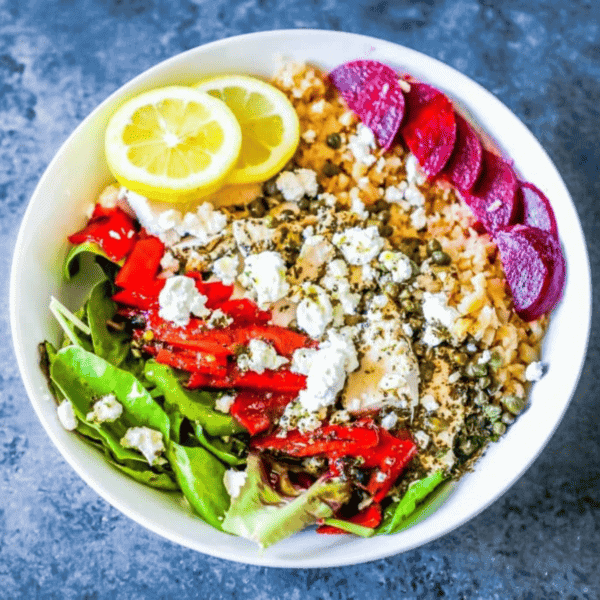 A colorful Greek chicken grain bowl with spinach, red peppers, beets, feta cheese, lemon, and a sprinkle of herbs.