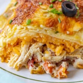 Close-up of a Chicken Enchilada casserole topped with chives and a black olive. Layers of shredded chicken, cheese, and diced vegetables are visible.