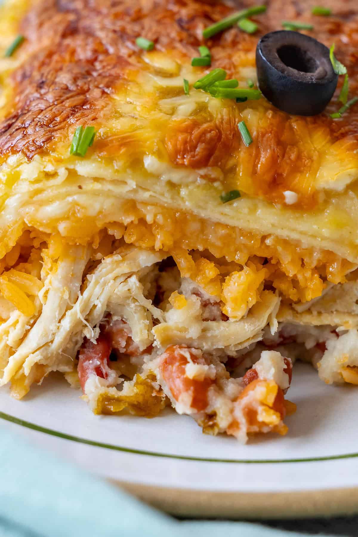 Close-up of a lasagna slice revealing layers of shredded chicken, melted cheese, and tomato bits reminiscent of a Chicken Enchilada Casserole, garnished with a black olive and chopped chives on top.