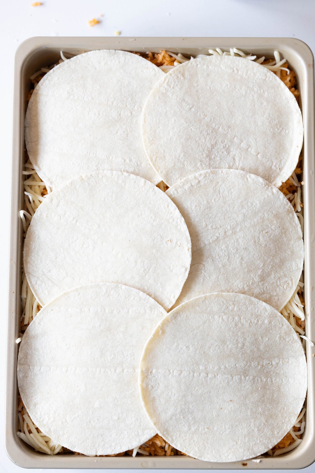A rectangular baking dish filled with shredded cheese and topped with six uncooked tortillas, perfect for creating an Easy Chicken Enchilada Pie Bake.