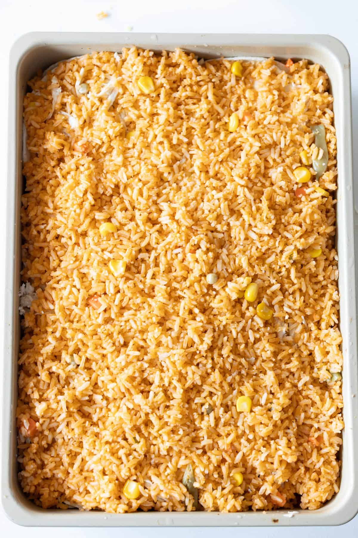 A rectangular baking dish filled with cooked orange-colored rice mixed with corn, resembling a delicious Layered Chicken Enchiladas casserole.
