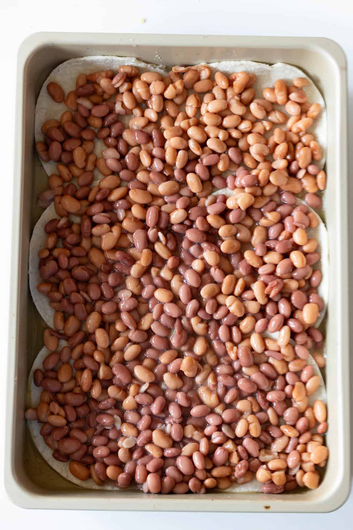 A baking tray filled with uncooked pinto beans spread evenly over a layer of tortillas, reminiscent of an Easy Chicken Enchilada Pie Bake preparation.