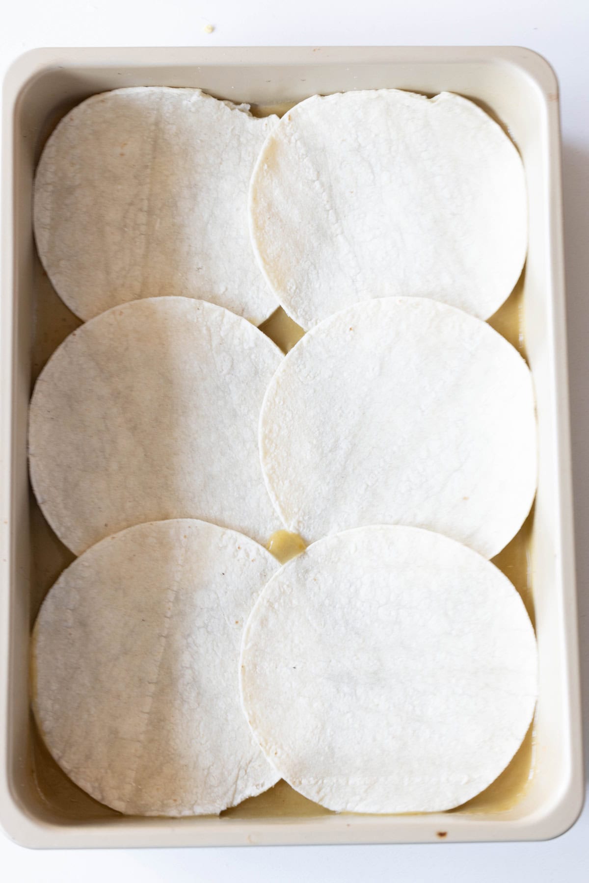 A baking dish with six circular white tortillas overlapping slightly, arranged in two rows, perfect for making a Green Chile Enchilada Casserole.