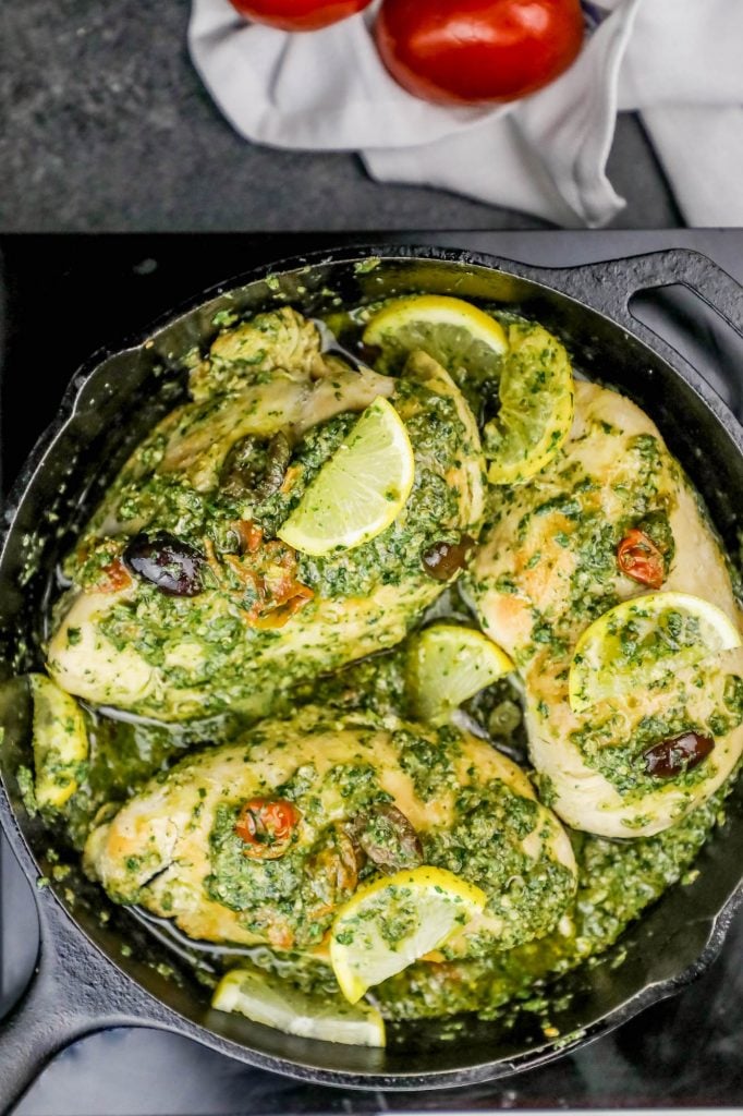 chicken covered in pesto and olives with a lemon slice on it in a cast iron pan