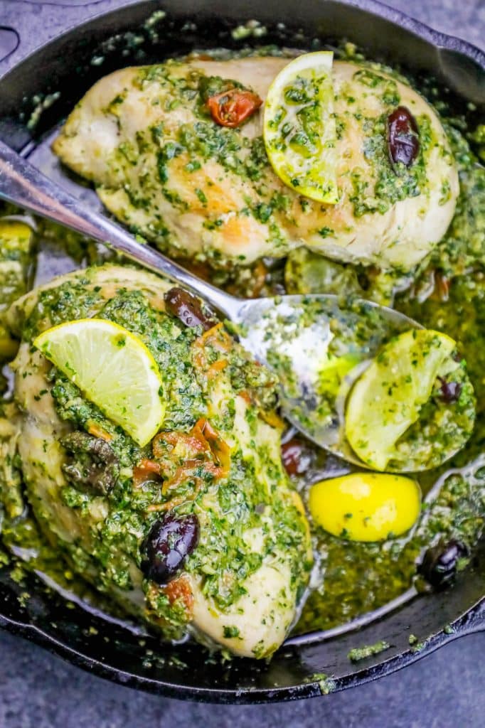 chicken covered in pesto and olives with a lemon slice on it in a cast iron pan with a metal spoon