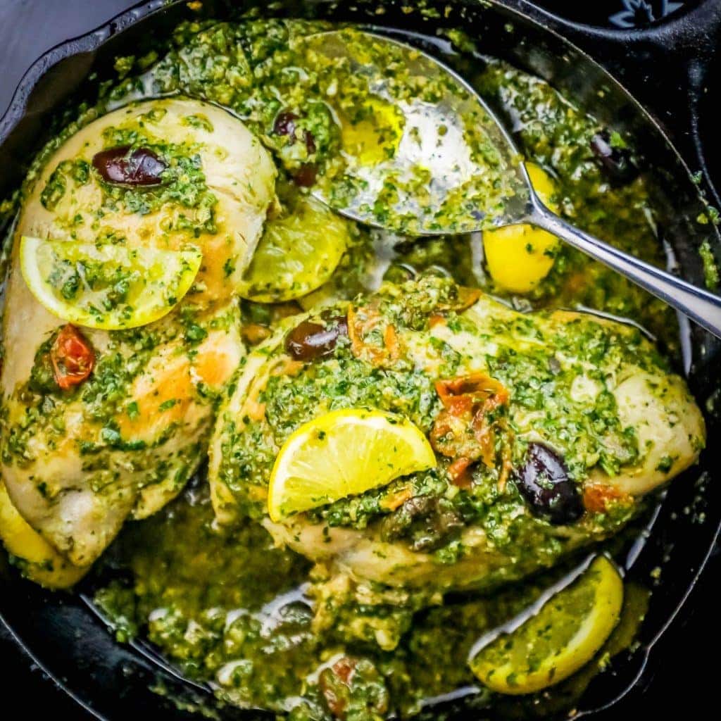 chicken covered in pesto and olives with a lemon slice on it in a cast iron skillet with a metal spoon