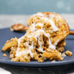 A serving of Cinnamon Roll Apple Crisp topped with a white icing glaze is presented on a dark plate.