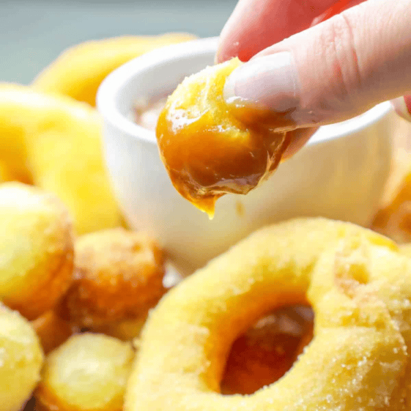 A person's fingers dipping a mini mulling spice donut into caramel sauce, with more doughnuts in the background.