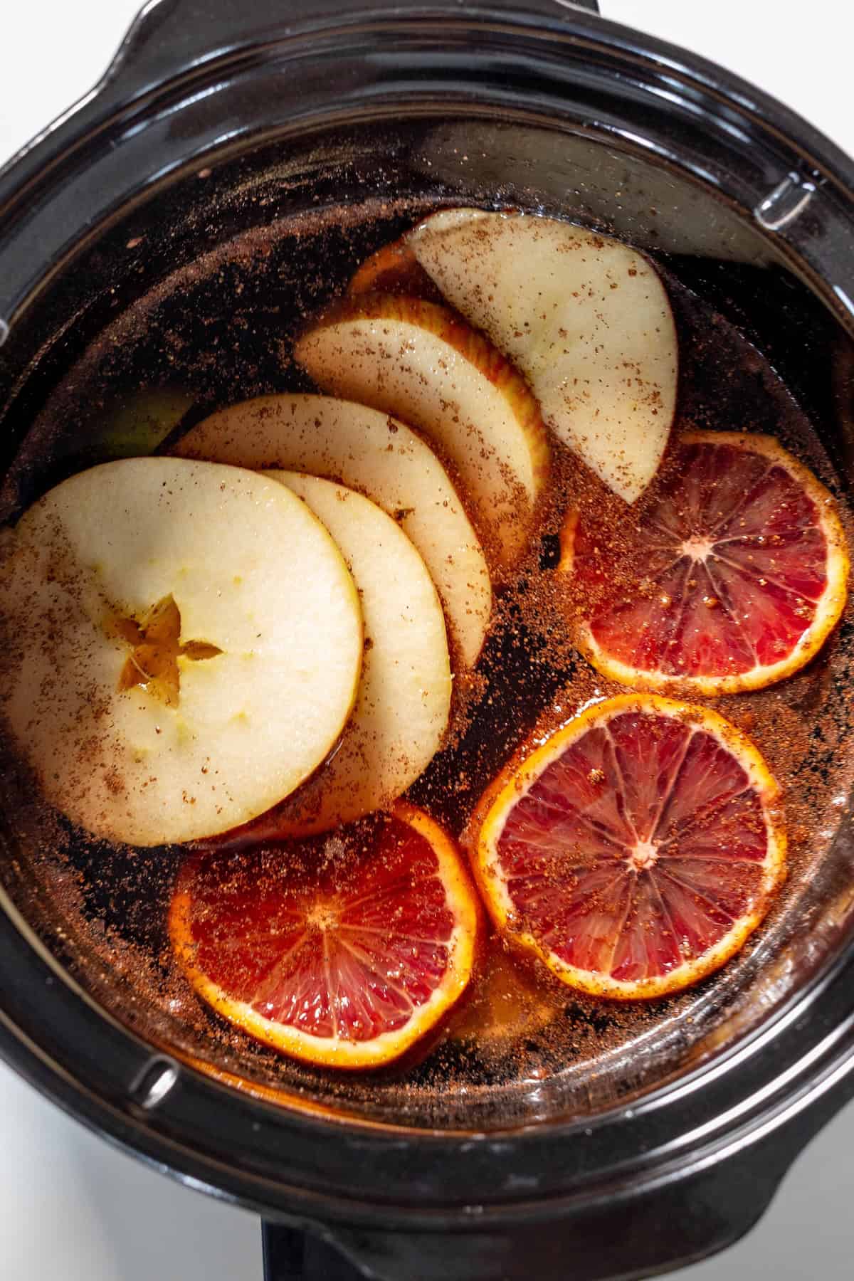 A slow cooker filled with sliced apples, blood oranges, spices, and liquid is prepped for the Best Mulled Cider Ever—perfect for cozy infusing or simmering.
