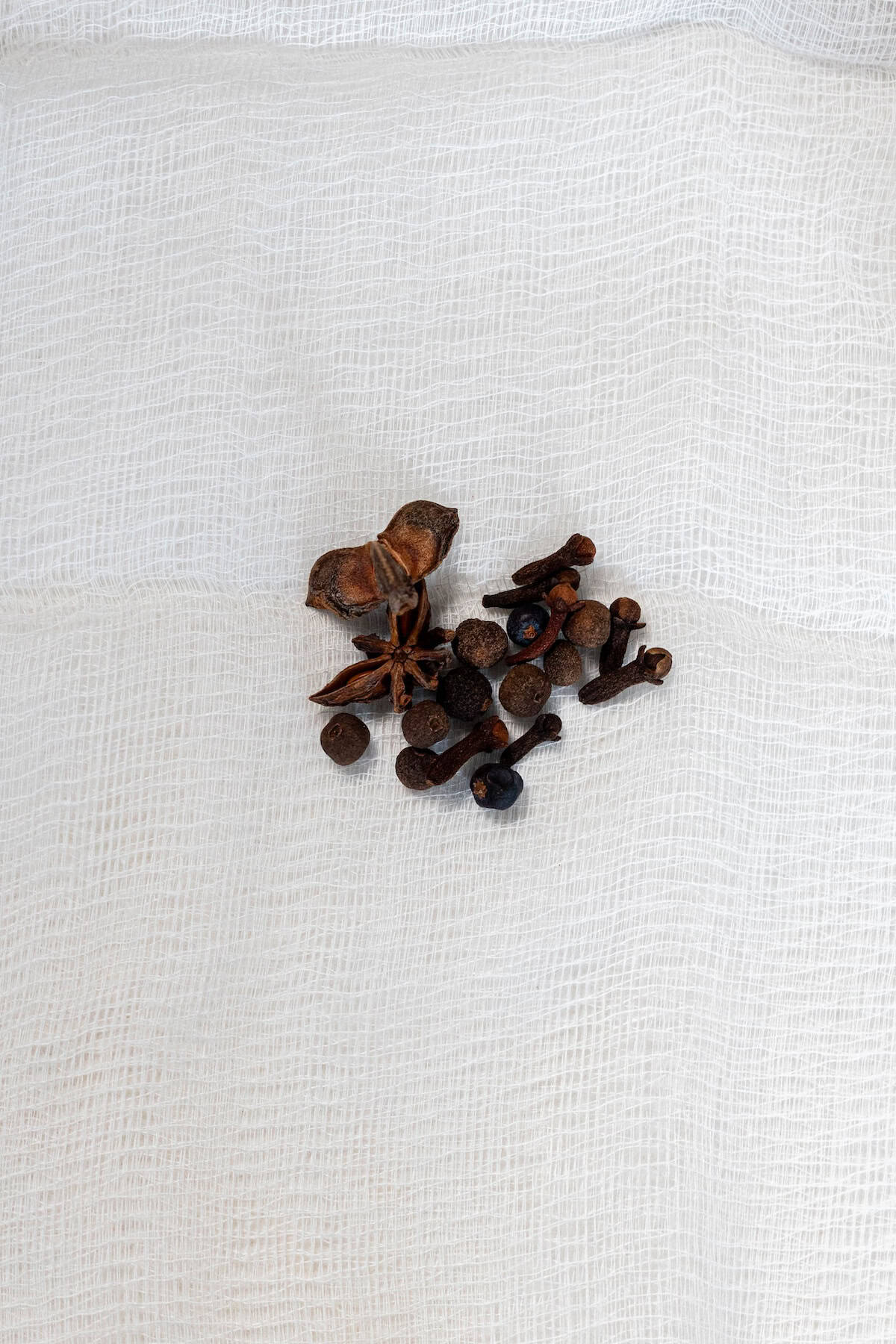 A small pile of assorted dried spices, perfect for the Best Mulled Cider Ever, features star anise, cloves, and peppercorns resting on a textured white cloth.