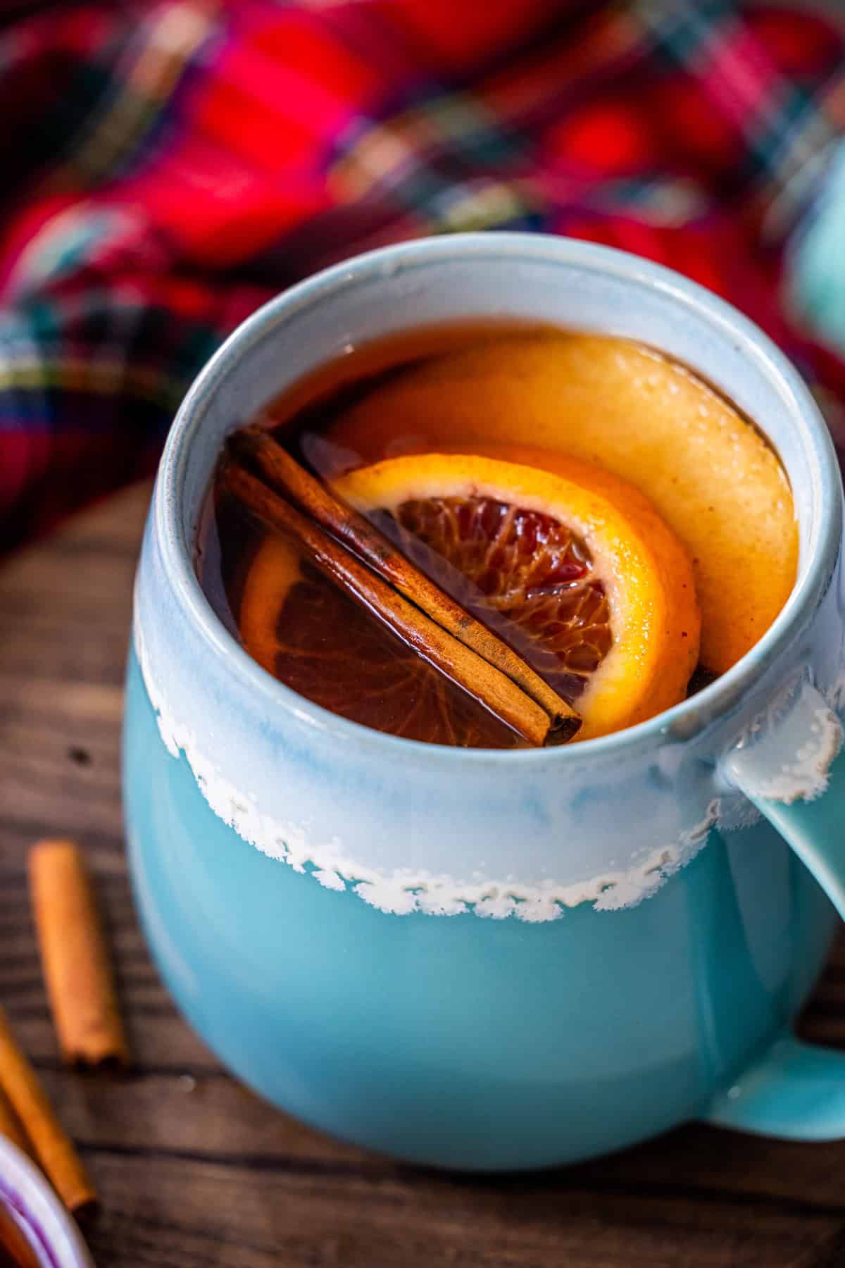 A blue mug filled with the Best Mulled Cider Ever, garnished with orange slices and a cinnamon stick, sits against a cozy plaid fabric background.