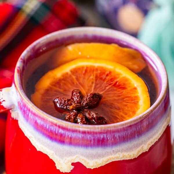 A red mug filled with the Best Mulled Cider Ever, garnished with a slice of orange and star anise, promises cozy warmth and festive flavor.