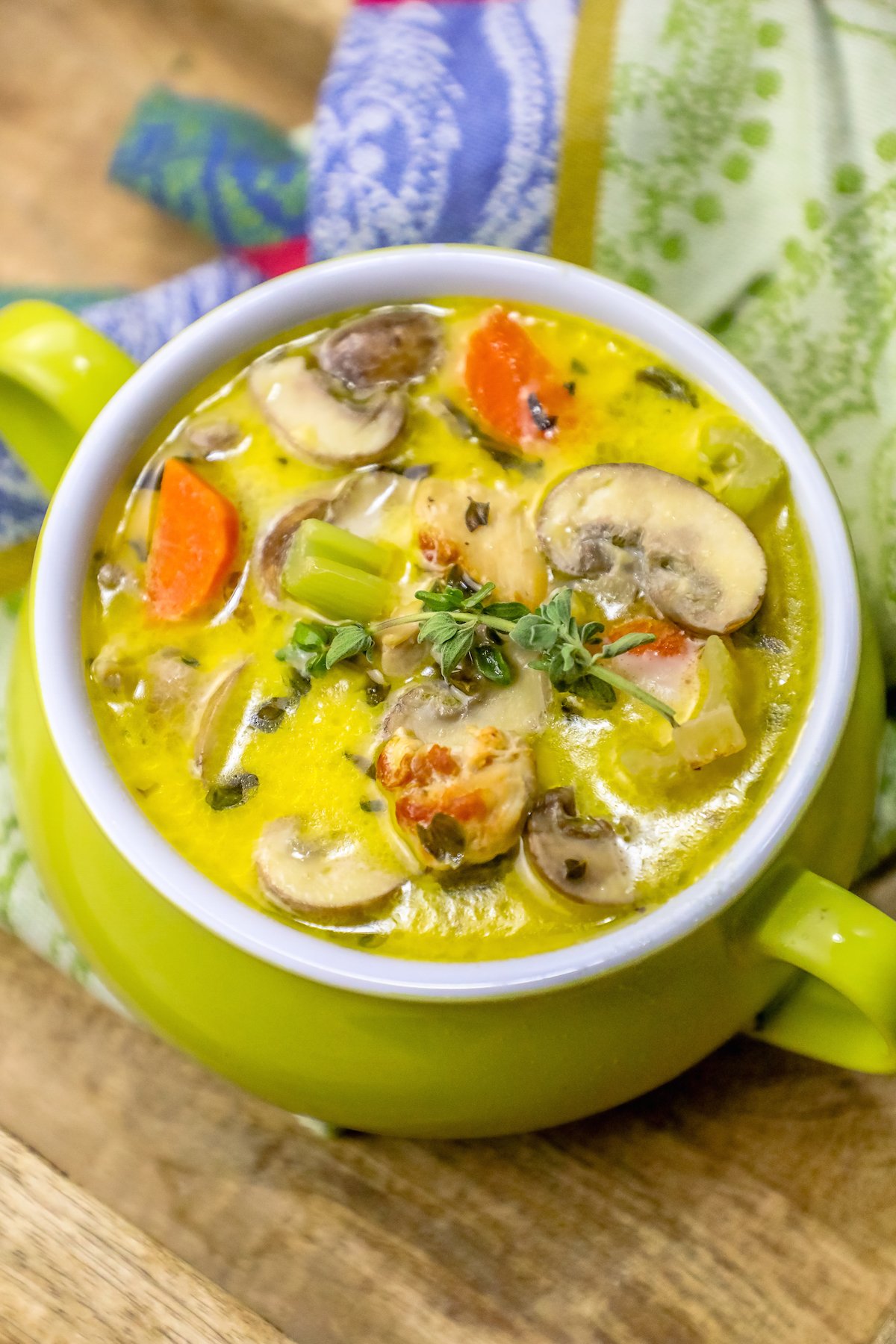 picture of mushroom and chicken soup with thyme on the top in a green pot with a colorful kitchen towel in the background. 