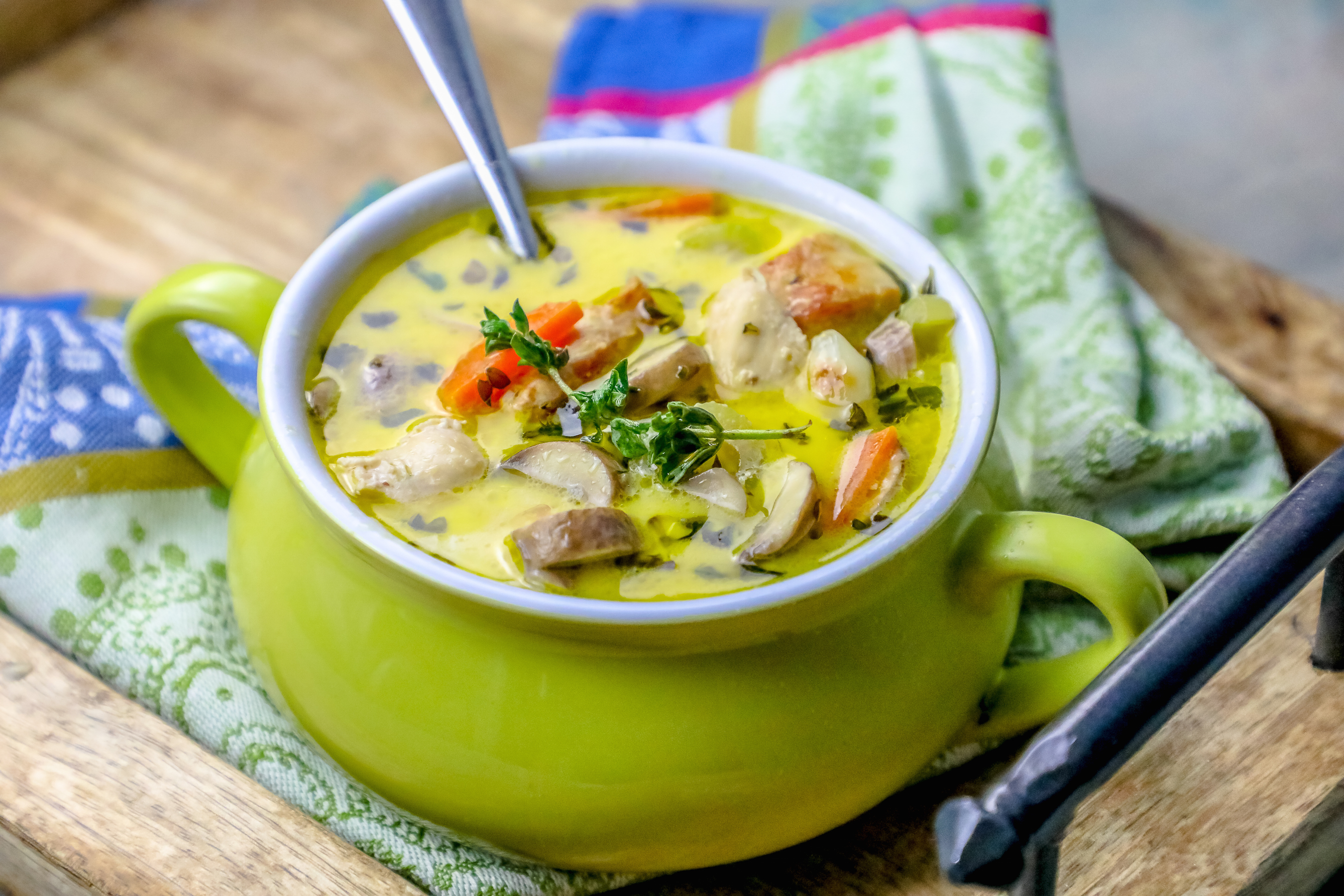 picture of mushroom and chicken soup with thyme on the top in a green pot with a colorful kitchen towel in the background. 