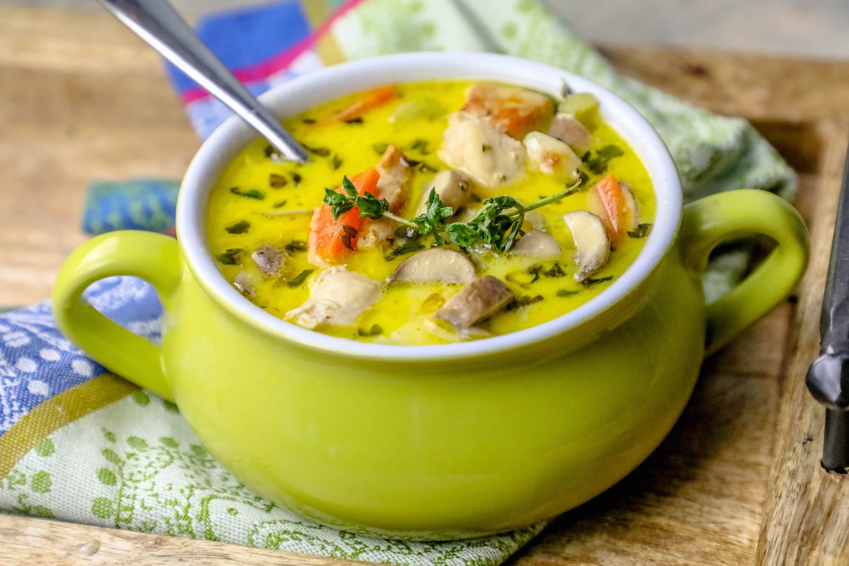 picture of mushroom and chicken soup with thyme on the top in a green pot with a colorful kitchen towel in the background. 