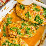 Three cooked chicken breasts are covered in a 3 Ingredient Nandos Peri Peri Chicken sauce, garnished with chopped parsley in a white baking dish. A wooden spoon rests on the edge of the dish.