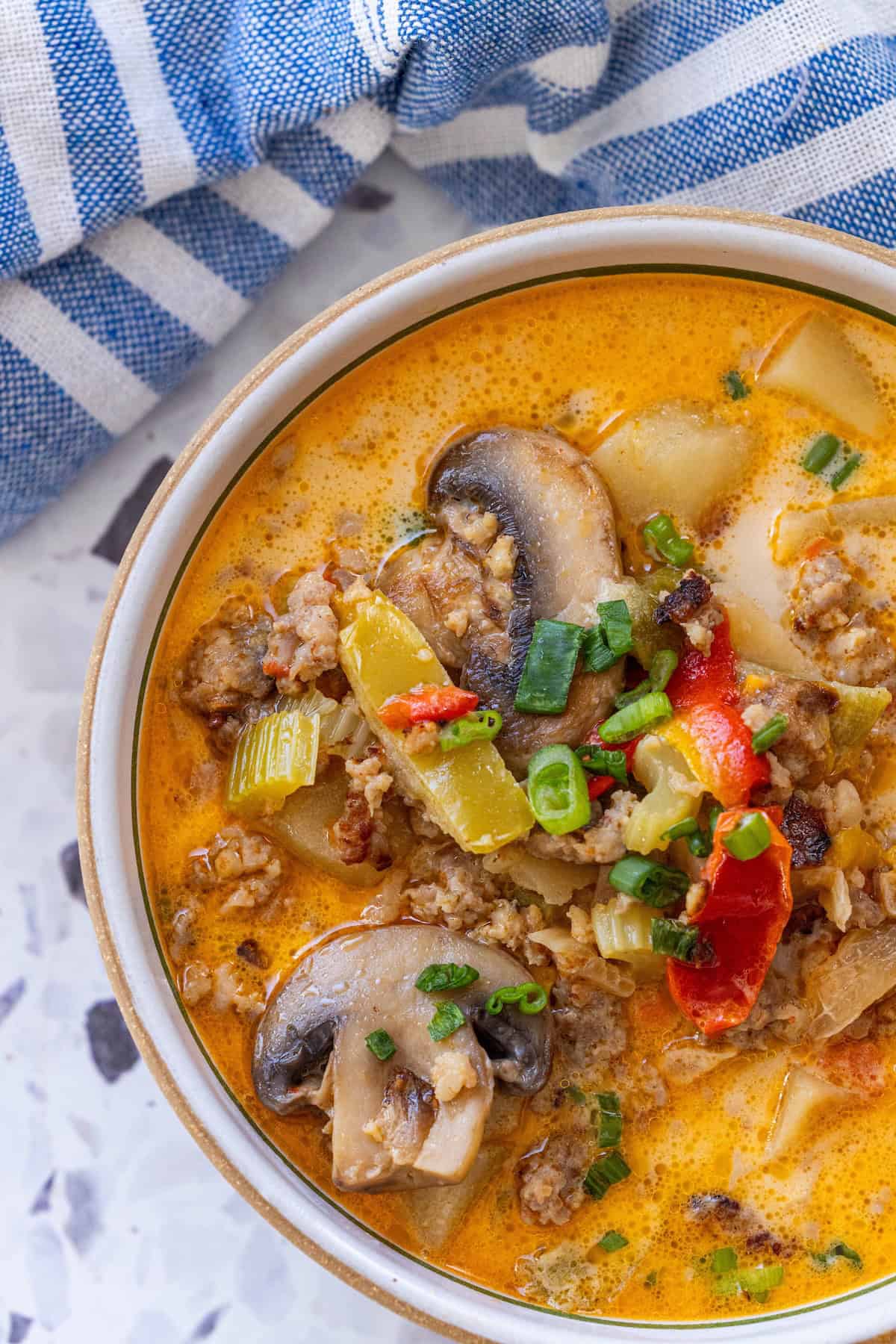 A bowl of creamy mushroom sausage chowder sits on a white surface next to a blue and white striped cloth—a delicious, one pot mushroom soup sausage recipe perfect for those following a keto diet.
