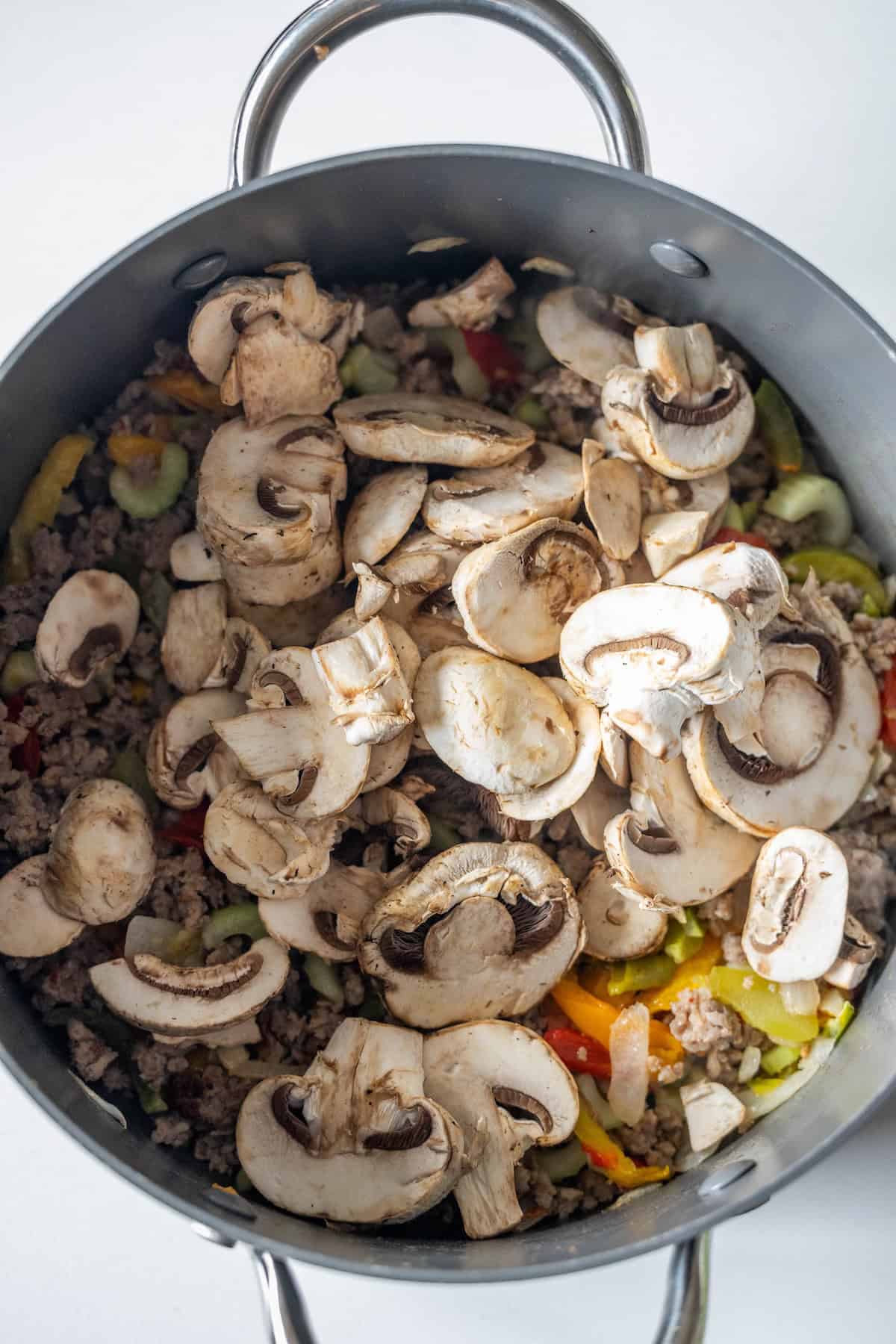 A pot filled with sliced mushrooms, ground meat, and chopped vegetables, perfect for an easy creamy mushroom and sausage soup recipe.