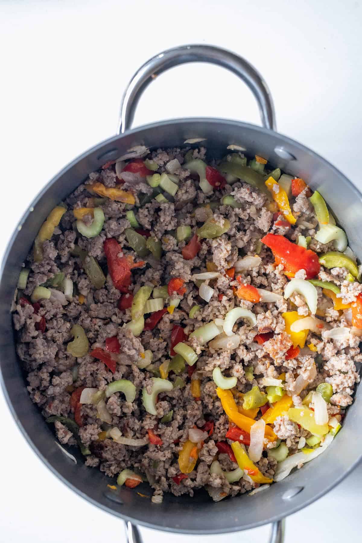 A pot filled with cooked ground meat, chopped celery, bell peppers, onions, and tomatoes on a white surface—perfect for a low carb mushroom sausage chowder.