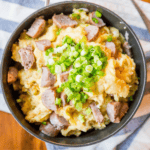 A bowl filled with mashed potatoes topped with sliced sausage and chopped green onions. Channeling a cheesy colcannon recipe, this hearty dish rests on a striped cloth, with a fork ready to dive in.