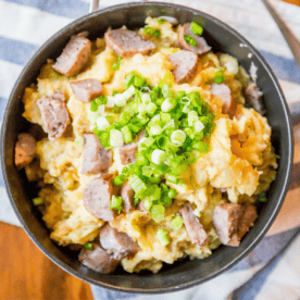 A bowl filled with mashed potatoes topped with sliced sausage and chopped green onions. Channeling a cheesy colcannon recipe, this hearty dish rests on a striped cloth, with a fork ready to dive in.