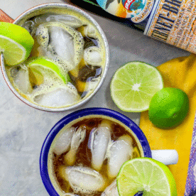 Two mugs filled with iced drinks garnished with lime slices, a cut lime, a whole lime, and a bottle are on a gray surface next to a yellow cloth—perfect for crafting an Italian Mule Cocktail with Fernet and Ginger Beer.