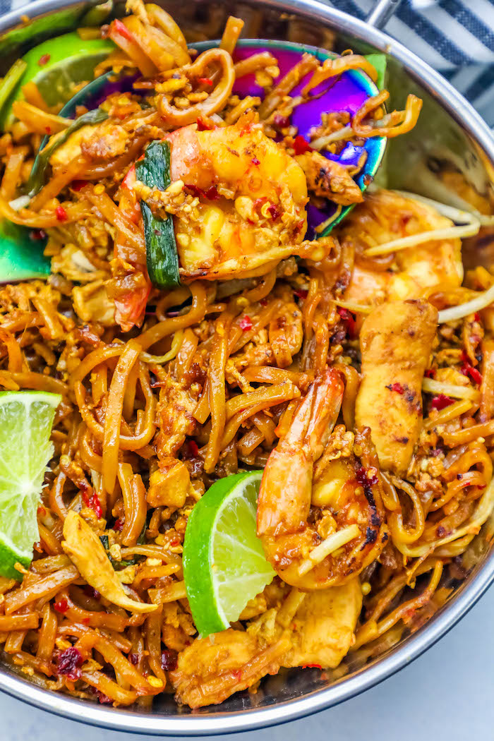 picture of pad thai with shrimp and limes in it and a spoon in it up close. 