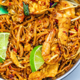 A close-up of stir-fried noodles with shrimp, chicken, vegetables, red chili, and lime wedges in a bowl—perfect for anyone looking to try The Best Easy Chicken and Shrimp Pad Thai Noodles Recipe for an authentic thai food recipe dinner.