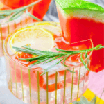 A glass of refreshing watermelon mocktail lemonade is served with a lemon wedge, a watermelon slice, and a sprig of rosemary for garnish.