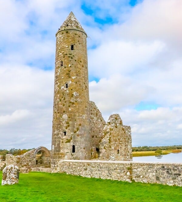 A stone tower stands in Clonmacnoise, Ireland, amidst the grassy fields of Ireland's Midlands with a view of a nearby body of water.