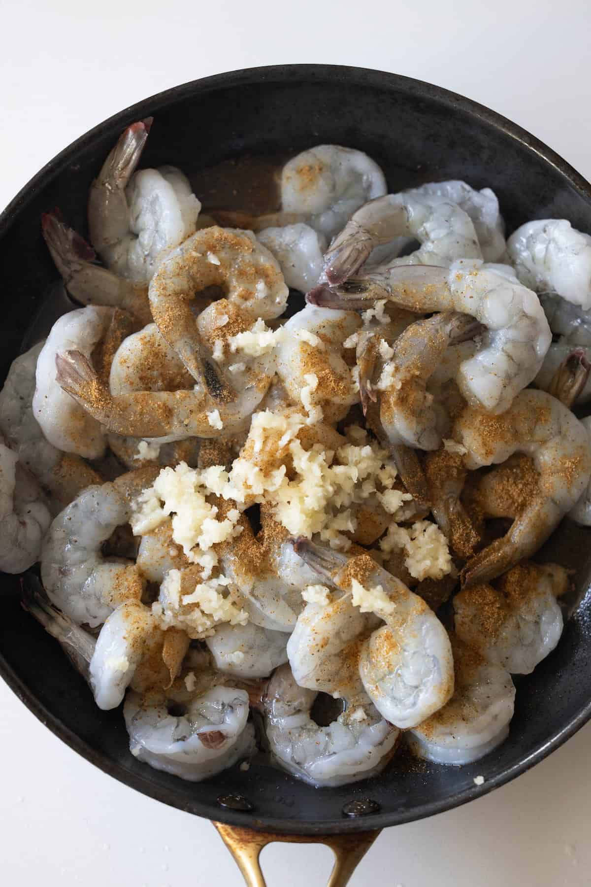 Raw shrimp in a black skillet topped with minced garlic and seasoning, ready to be cooked for cilantro lime shrimp&mdash;perfect if you're learning how to cook shrimp that doesn&rsquo;t taste fishy.