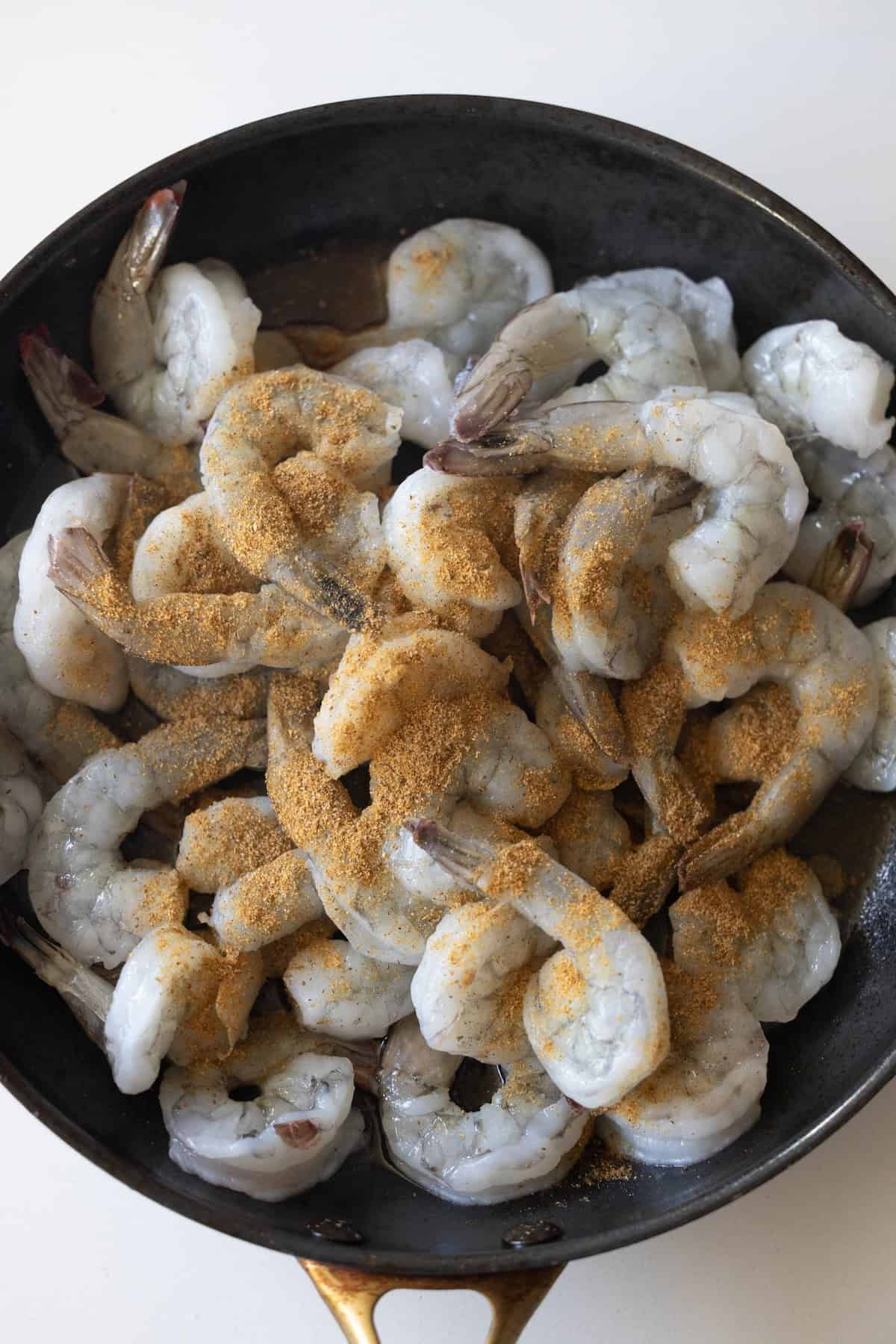 Raw shrimp in a black skillet, sprinkled with a layer of seasoning powder, ready to become an Easy Cilantro Lime Shrimp dish. Learn how to cook shrimp perfectly every time.