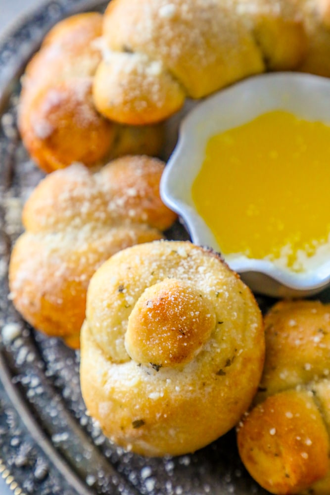 baked garlic knots dusted with parmesan and herbs next to a cup of melted butter on a platter