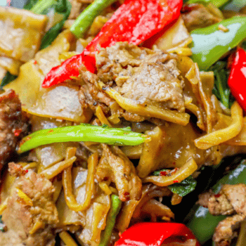 A close-up of beef drunken noodles showcases stir-fried noodles with sliced beef, red and green bell peppers, and vibrant leafy greens. (Don't worry—drunken noodles don't actually contain alcohol!)