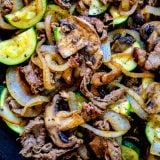 A skillet filled with zucchini and onions cooked alongside thin sliced top sirloin for a delicious dinner.