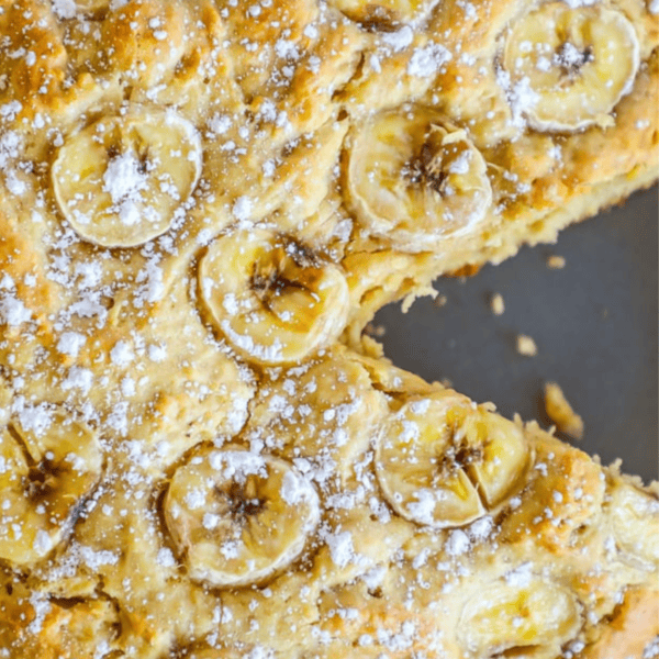 Apple banana bread topped with sliced bananas and dusted with powdered sugar sits on a grey surface, with a tempting slice missing.