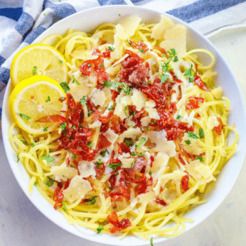 A bowl of creamy lemon linguine recipe topped with sun-dried tomatoes, shaved parmesan, chopped parsley, and two lemon slices, on a white surface with a striped cloth nearby.
