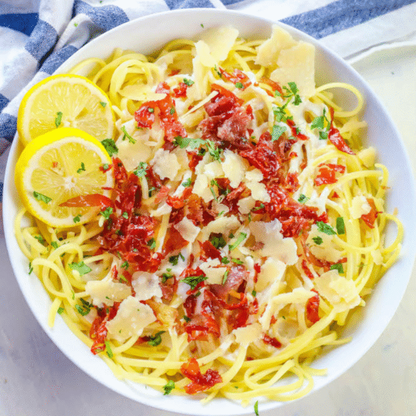 A bowl of creamy lemon linguine recipe topped with sun-dried tomatoes, shaved parmesan, chopped parsley, and two lemon slices, on a white surface with a striped cloth nearby.