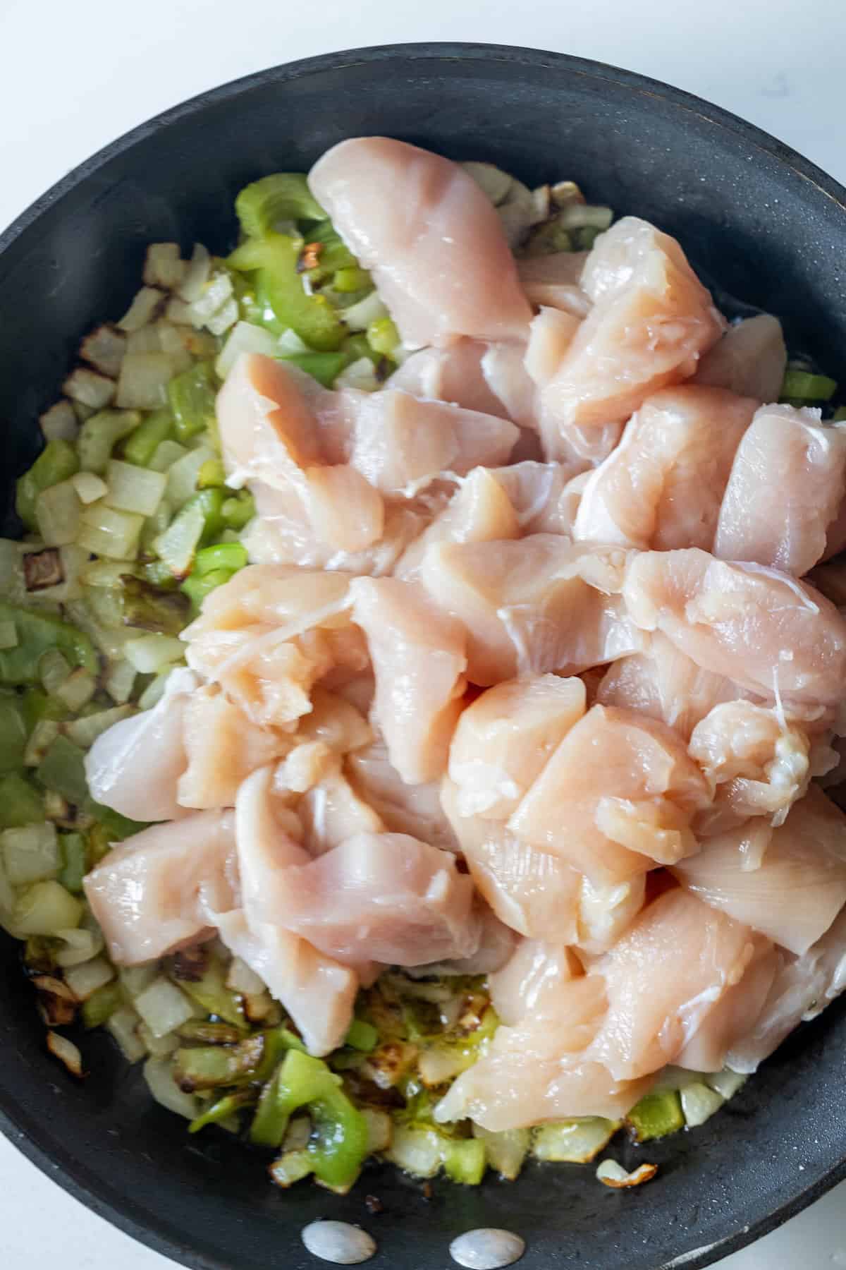 Raw diced chicken rests atop sautéed onions and green bell peppers in a black skillet, perfect for a quick 20 minute Italian chicken and vegetables skillet.