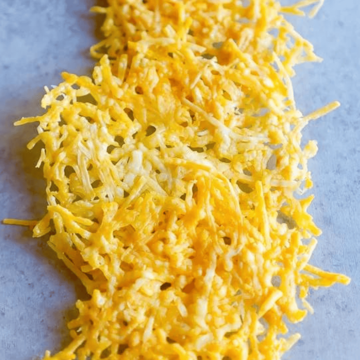A pile of finely shredded yellow and white cheese is spread out on a flat grey surface, perfect for making an easy baked Parmesan crisps recipe.