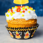A pumpkin cupcake with white frosting and red, yellow, and orange sprinkles sits perfectly in a Halloween-themed wrapper.