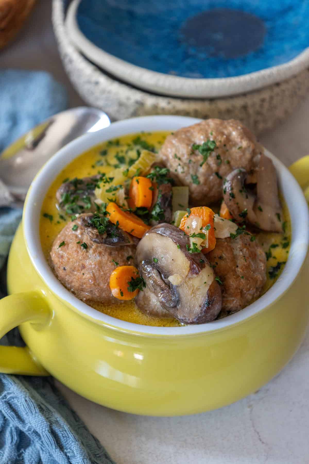 A yellow bowl brimming with a one pot meatball soup recipe, featuring tender meatballs, mushrooms, carrots, and herbs in a creamy sauce. It sits invitingly on the table beside a blue towel and ceramic bowls.