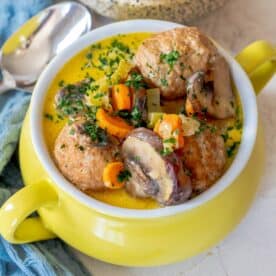 A delicious yellow bowl of keto meatball soup features tender meatballs, carrots, mushrooms, and herbs. A silver spoon rests beside the bowl on a blue cloth, inviting you to savor this comforting one pot meal.