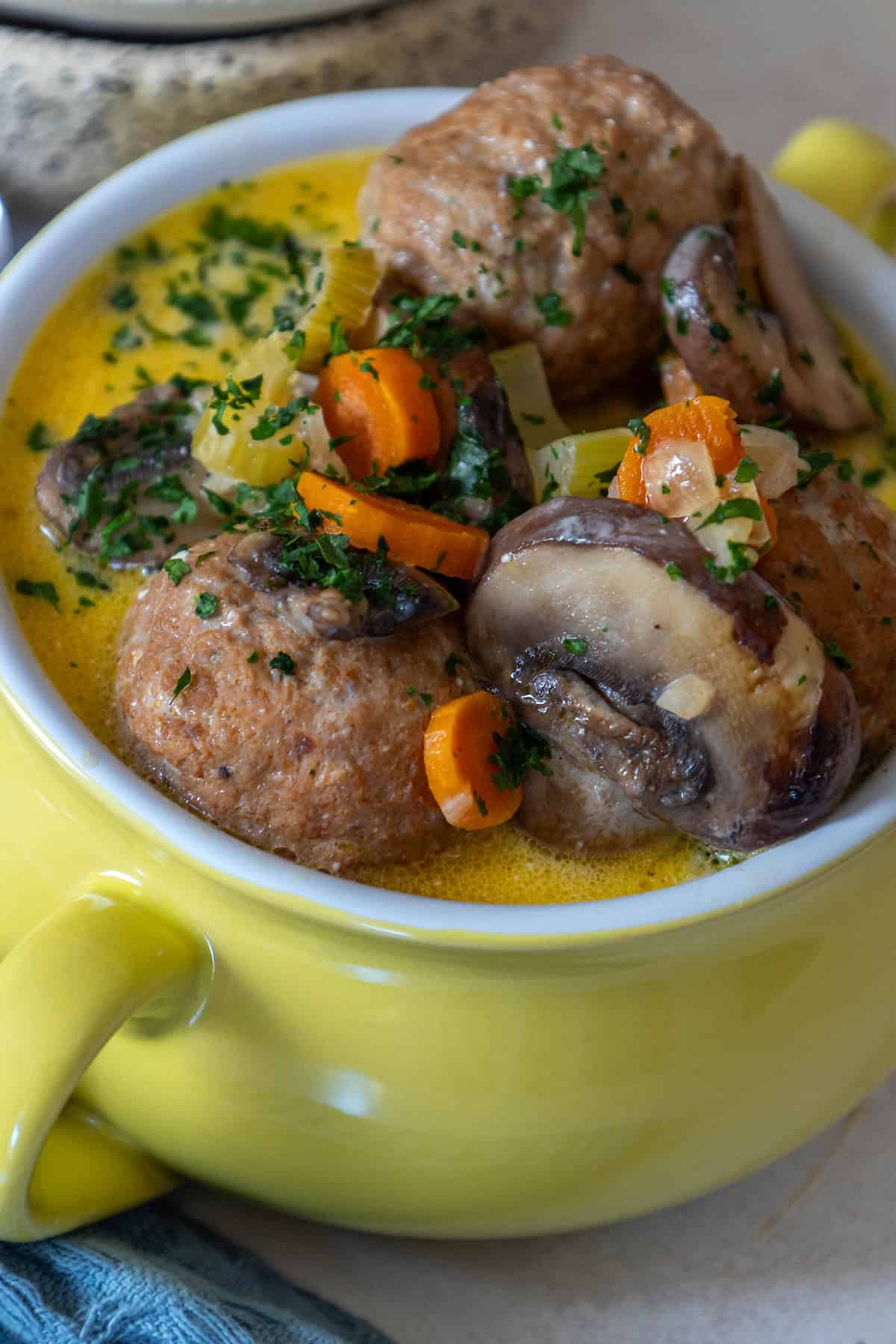 Close-up of a yellow bowl with keto meatball soup, featuring savory mushrooms, carrots, and herbs in a creamy sauce.