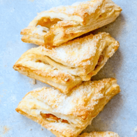 Three golden, sugar-dusted puff pastry turnovers with visible fruit filling are stacked on a light gray surface, perfectly showcasing this easy apple turnovers recipe.